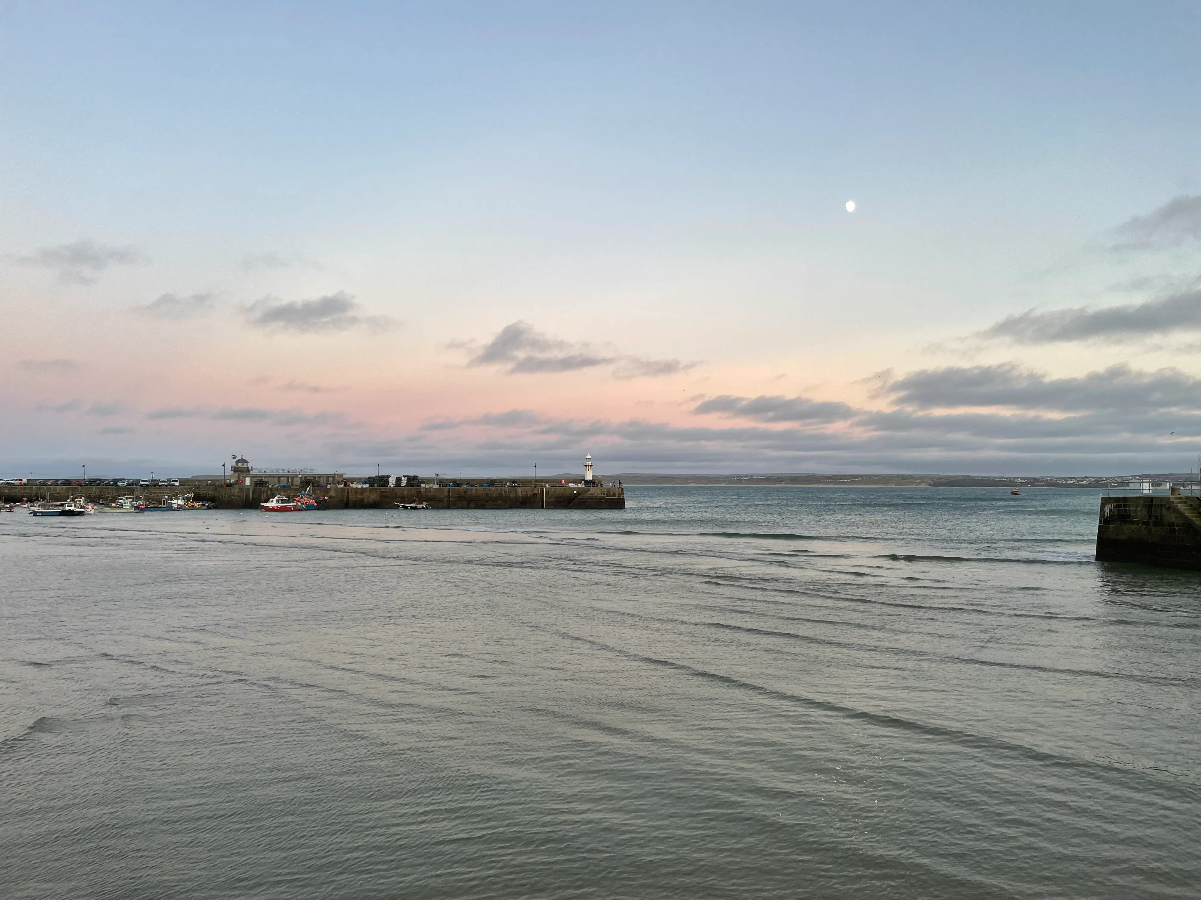St Ives harbour at dusk — calm water stretching towards the pier and lighthouse, the moon hanging in a soft pink sky
